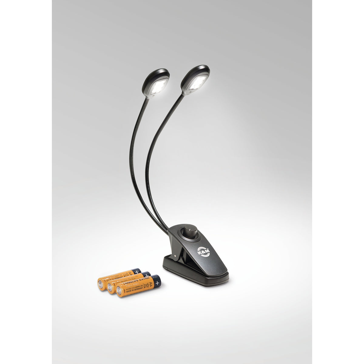 12271 Twin Head Pro music stand light with both LED heads illuminated on grey backdrop, clip base with K&M logo and dimmer knob, three AA batteries beside it, K&M