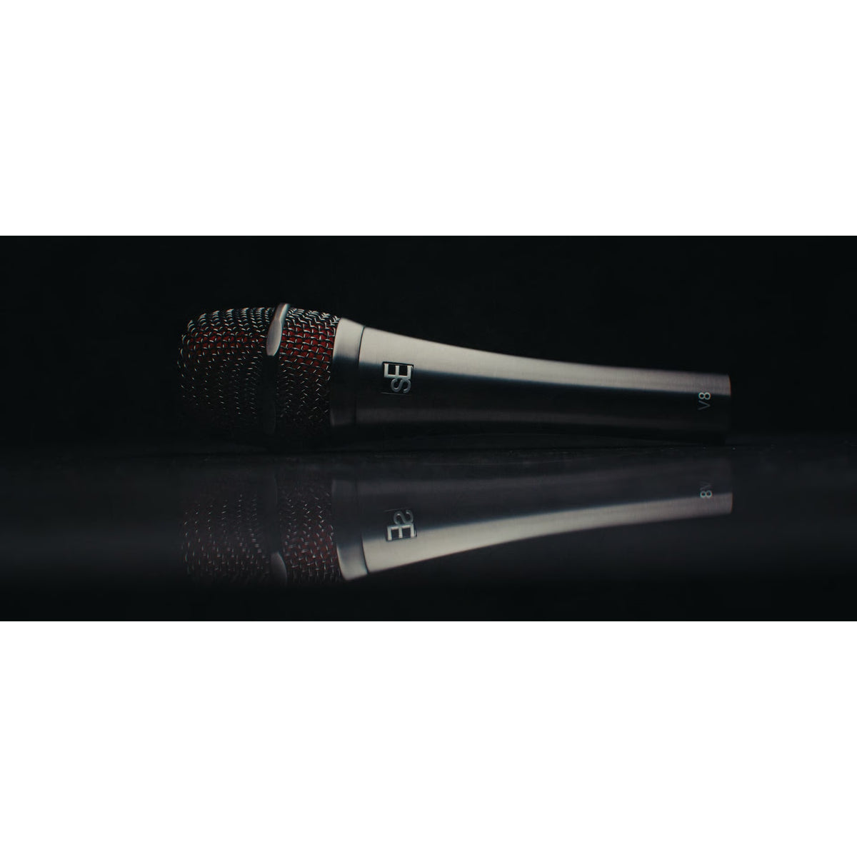 V8 handheld vocal microphone lying on a reflective black surface in a moody studio shot, showing red-lined grille, sE logo and V8 marking, sE Electronics