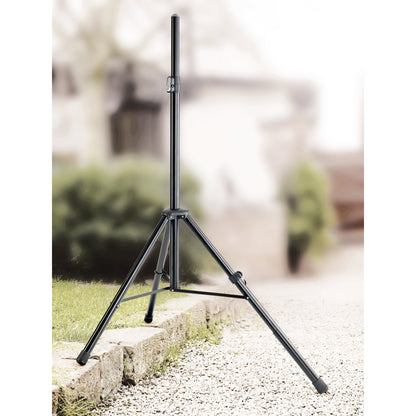 Black tripod speaker stand set up outdoors on a stone curb and gravel, one leg extended to level the stand on uneven ground, K&M