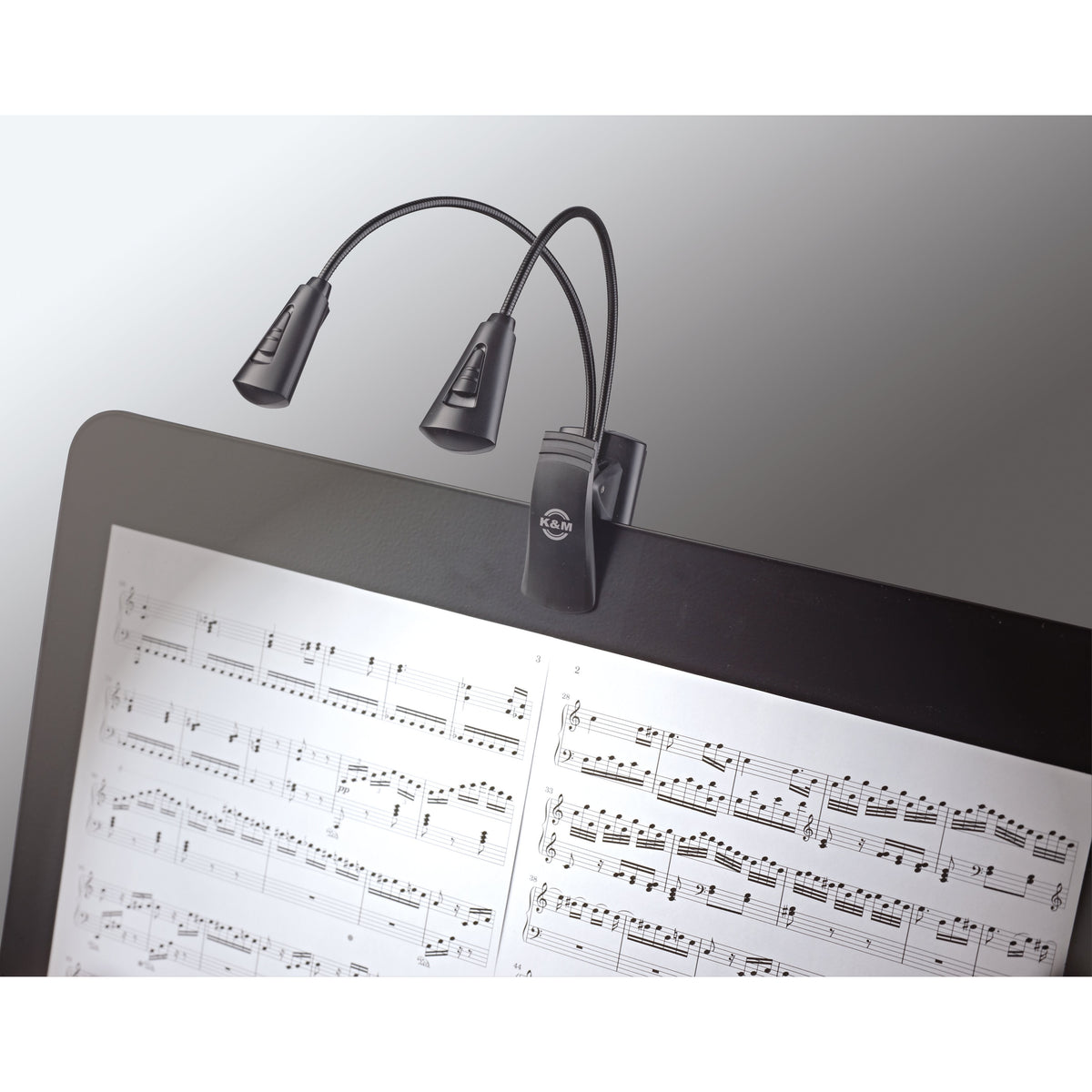 12244 dual-head LED FlexLight in use clipped to a music stand, lamp heads angled to illuminate open sheet music, K&M