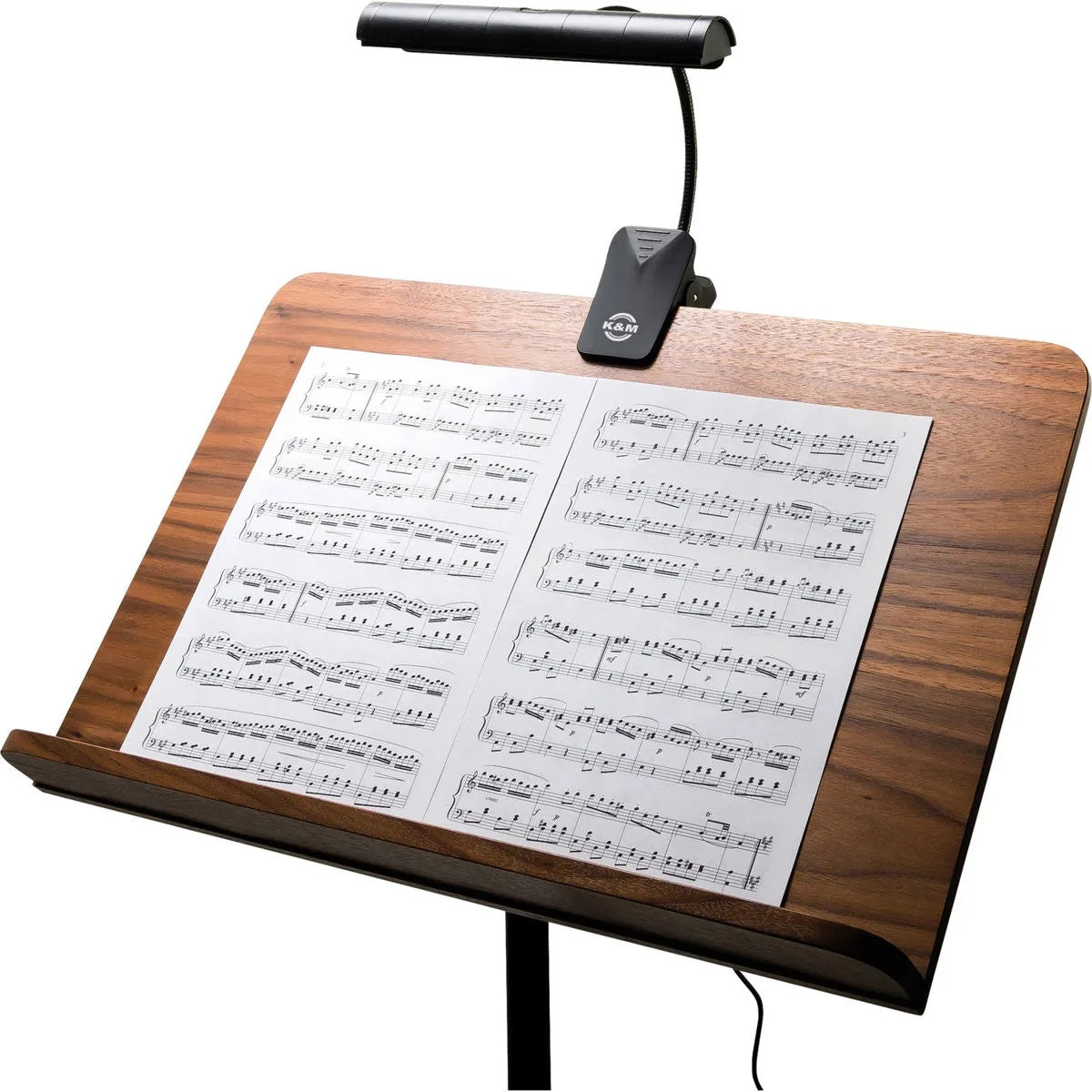 12287 Orchestra LED music stand light in use, clipped to the top of a wooden music desk and illuminating open sheet music, K&M