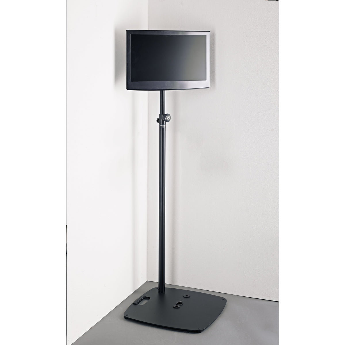 19610 stand adapter in use holding a flat-screen monitor on a black extension tube and weighted floor base in a room corner, K&M